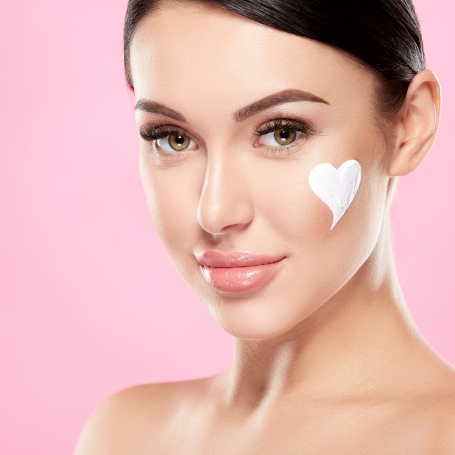 Cute model with brown hair fixed behind, clean fresh skin, big eyes and naked shoulders posing at pink studio background and looking at camera, close up, using face cream.